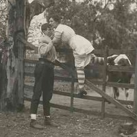 Image: Theatrical still of a young woman in 1920s attire being helped over a high gate by a young man. Behind the gate is a cow, suggesting the woman’s narrow escape from danger