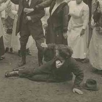 Image: A theatrical still of a group of people looking on with concern as two young women attempt to restrain a man from hitting another man who has fallen to the ground in a fight. In the background is a country homestead
