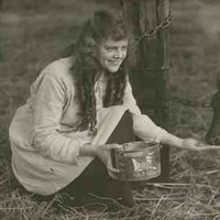 Image: A theatrical still of a young woman squatting beside a tethered calf as she feeds it from a tin. The label on the tin has a brand name for Country Stores, 290 Rundle Street, Adelaide