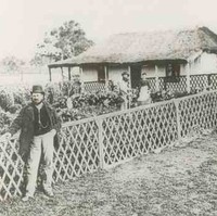 Image: A man in a bowler hat leans against a fence surrounding a garden. A small cottage connected to the garden is in the background