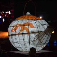 Image: A large white and orange paper lantern in the shape of a teapot glows in the dark as it is carried by a group of people in front of a stage lighting rig illuminated in pink and a banner reading moon lantern festival. 
