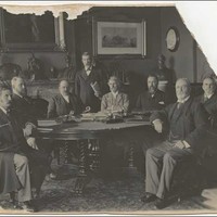 Image: photograph of seven men sitting around a table with an eighth standing behind. The photograph is torn in the top right corner. 