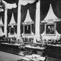 Image: a group of men sit at ornate wooden desks in a room lined with white columns and decorated with portraits, dark coloured curtains and swathes of dark fabric hanging from the ceiling. 