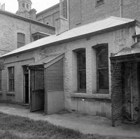 Image: A low, rectangular stone and brick building surrounded on three sides by much taller brick and stone buildings. The back door of the building features a wooden latticework awning