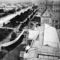 Image: Black and white photograph of a tree lined boulevard, as viewed from the roof of a building. The buildings lining one side of the road are grand stone constructions and are surrounded by formal gardens.