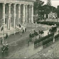 Image: lines of men in military uniforms plus others on horseback gather outside a large stone building with wide steps leading to its entrance and decorative columns along the entire facade. 