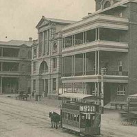 Image: a horse drawn tram travels down a wide dirt city street which is lined with a series of three and four storey buildings, many with balconies and verandahs, including a theatre with a large arched entrance and column decoration on the upper floors. 