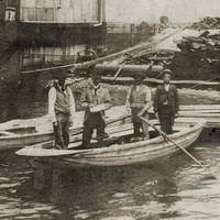 Image: Four men stand in a wooden row-boat next to landing steps on a riverbank. Two other empty row-boats are moored nearby, and a large building is visible on the shoreline in the immediate background 