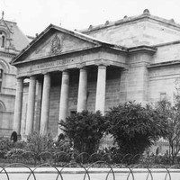 Image: A large, mostly windowless sandstone building fronted by a portico with six columns