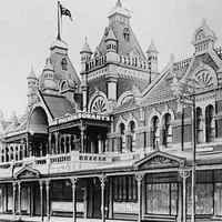 Image: Black and white image of store front building with tiled roof, turreted towers and byzantine details. Sign reads "Grants Coffee Palace"