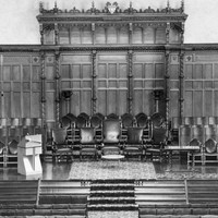 Image: a low stage, with the wall behind covered in decorative wooden panelling, supports rows of chairs, a small table and a lecturn. The stage also has further rows of chairs facing it. 