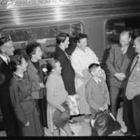 Image: Six people, both adults and children stand in front of a train on a station platform. They are laughing and conversing. 
