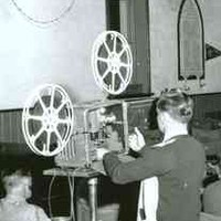 Image: A young man operates a film projector in a large church hall filled with seated people of all ages
