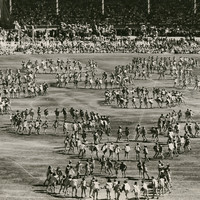Image: An oval and grandstands filled with people, children dance on the oval in circular formations