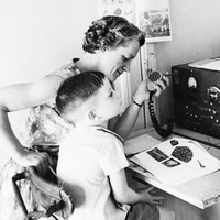 Image: Two adult women and a young boy sit in front of a large two-way radio receiver. One woman holds the radio's transmitter in her hand. The boy has an open textbook in front of him