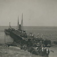 Image: A wooden jetty jutting out into the ocean with several horses and wagons and two large sail boats docked at the side