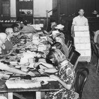 Image: A group of women sit on either side of a long table arranging paper documents, a man walks by with a trolley stacked with papers. 