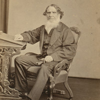 Image: A man wearing a suit sitting in a chair with one hand resting on a small pedastal