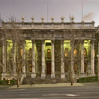 Image: a large stone building with two storey high columns along the entire facade, and wide stone steps leading up to its front entrance, is lit up with green lights at dusk.