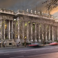 Image: A large stone building with two storey high columns lining its front is lit up by green lights at dusk.