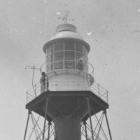 Image: A metal lighthouse stands on a desolate island. Two men stand in the lantern room at the top of the lighthouse, while a third sits outside the keepers’ quarters at its base. A horse and cart stands next to the seated man