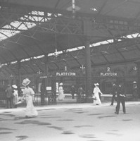 Image: the interior of a railway station with at least three platforms visible. The curved roof features skylights and is supported by metal beams. In the foreground are a number of men and women in early 20th century clothing.