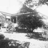 Image: A large stone house bordered on one side by a well-tended garden. A woman stands on the verandah of the house 