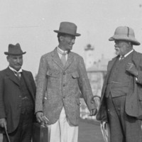 A man in a suit and hat is greeted by other men on a jetty. A coastal town is visible in the background