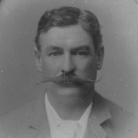 Image: A photographic head-and-shoulders portrait of a man in suit and tie sporting a large handlebar moustache. The frame surrounding the portrait identifies the man as Richard Chaffey Baker