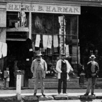 Image: three men stand outside a drapery shop in part of a two storey terrace building. A figurine of a woman holding a trident can be seen above the door to the shop.  