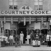Image: A group of men and children stand outside a two storey terrace building the ground floor of which is serving as a drapery store with fabric sitting in baskets on the street, coats hanging along the storefront and hats in the window. 