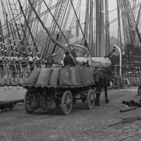 Image: Horse-drawn carts containing numerous large sacks of flour travel along a wharf next to a large, three-masted sailing ship
