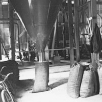 Image: The interior of a large warehouse with belt-driven machinery. Full gunny sacks are arranged near the machinery, and one sack is positioned beneath a large funnel