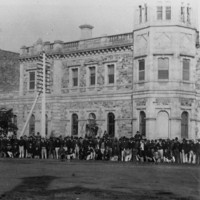 Image: A large group of men dressed in late nineteenth century attire pose for a photograph in front of a large, two-storey stone building with a corner octagonal tower. A dirt street is visible between the photographer and the group of men