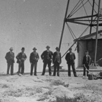 Image: A lighthouse stands at the top of a hill on a desolate island. A group of men in early Edwardian apparel pose for a photograph next to and in front of the lighthouse tower