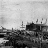 Image: A shallow creek contains two boats, one upturned. A small group of Aboriginal people sit in the foreground. On the right is a busy street with horse drawn wagons taking bales to be loaded onto ships in the background