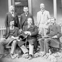 Image: black and white photograph of six men, three seated and three standing in front of a building. Kingston is seated in the front middle and wears a flower in his lapel.
