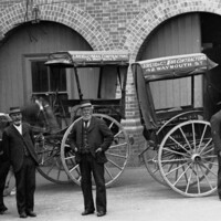 Image: Five men in uniforms stand in front of three horse-drawn carts in a building courtyard. Painted on the side of the carts are the words ‘J. Reid & Co. Mail Contractors, 42 Waymouth St.’ 