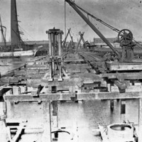 Image: The supporting framework of a bridge under construction extends across a river. Sailing ships, a wharf, and a large brick smokestack are visible in the image background