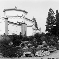 Image: An ornate conservatory building sits within a densely planted garden in full bloom with a winding gravel path in the foreground.  