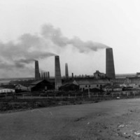 Image: Several large brick chimneys are flanked by a number of buildings. A vague coastline is visible in the background
