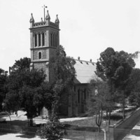 Image: A large, stone church surrounded by trees stands on the corner formed by two streets. A man in a brimmed hat stands in front of the churchyard