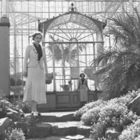 Image: Two women in pale skirts and jackets over dark blouses stand apart from each other outside a large glass conservatory building in this posed black and white photograph from the 1930s.