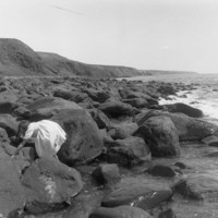 Image: View of a rocky coastline with a young girl clambering over rocks and cliffs in the background 