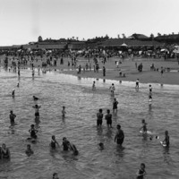 Image: View of beach with people bathing in the water and crowds on sand in the background