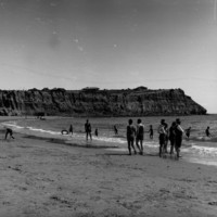 Image: Beach scene with people playing on the sand and in the water with rugged cliffs in the background