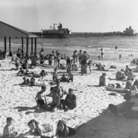 Image: People sunbathing on a beach with a jetty in the background.