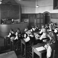 Image: Girls in black pinafores over white blouses sit at wooden desks with open books facing a blackboard. Under the blackboard, behind a large desk on a raised platform sits a woman, their teacher, also with an open book.