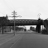 Image: two flights of stairs lead up to a metal bridge which passes over a wide paved road upon which an electric tram travels