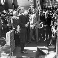 Image: a large crowd gathers around a man in a 1920s era dark suit who stands on am small stage with a carved foundation stone.